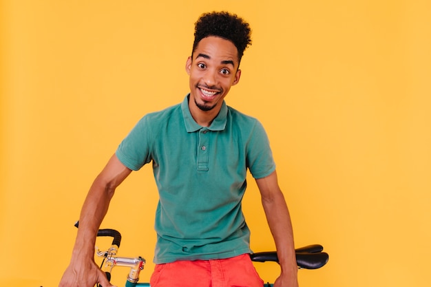 Cycliste en t-shirt vert posant avec un sourire surpris. Portrait de mec aux cheveux noirs étonné assis près de vélo.