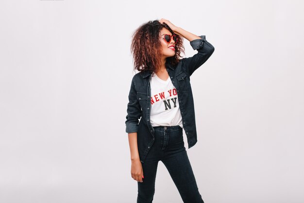 Dame africaine à la mode en tenue de denim noir en levant tout en posant. Photo intérieure d'une fille spectaculaire dans des lunettes de soleil drôles isolées.