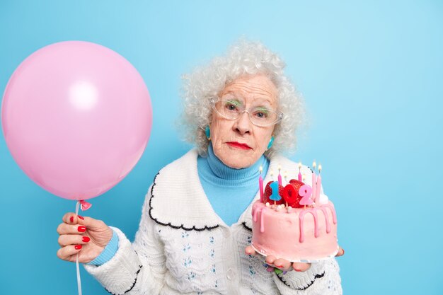Une dame âgée à la mode sérieuse regarde directement, célèbre des poses d'anniversaire avec un gâteau de fête et un ballon gonflé en pension