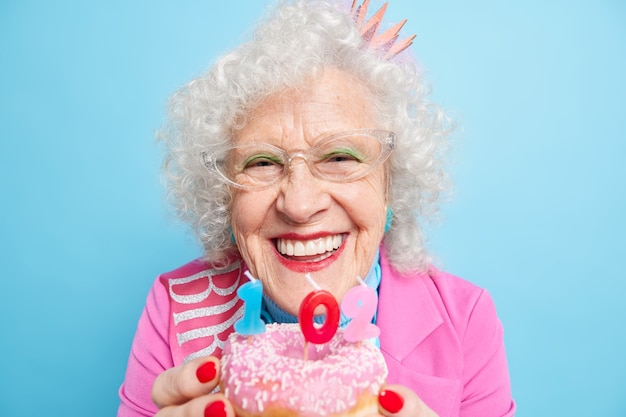 Photo gratuite une dame âgée positive sourit largement a une humeur festive souffle des bougies sur un beignet fait un vœu pour son 102e anniversaire a l'air parfait a un maquillage lumineux porte des vêtements élégants et élégants