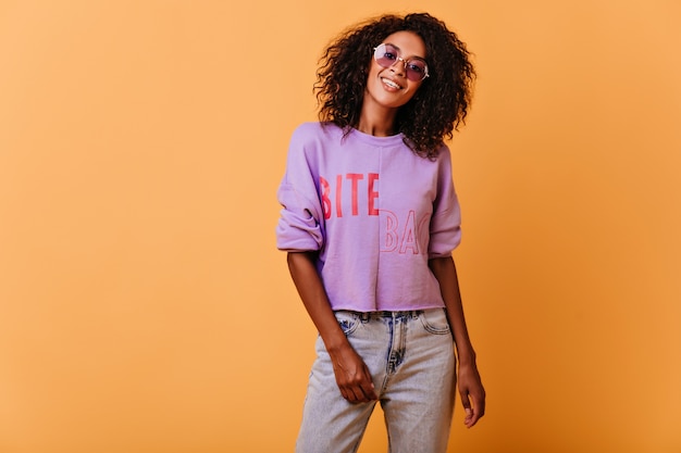 Dame agréable à la mode dans des lunettes de soleil debout sur le jaune. Jolie femme africaine exprimant des émotions sincères.