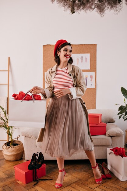 Dame au chapeau rouge et manteau pose avec sac à provisions. Belle fille aux lèvres brillantes en jupe longue, en talons hauts et sourire de pull rayé.