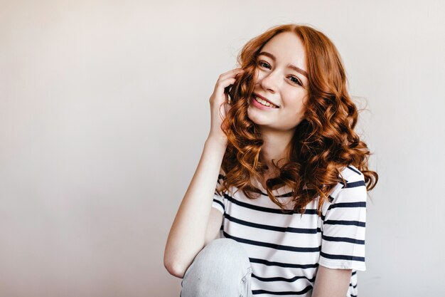 Dame au gingembre aux yeux sombres assis et riant. Tir intérieur d'une magnifique fille caucasienne en t-shirt rayé.