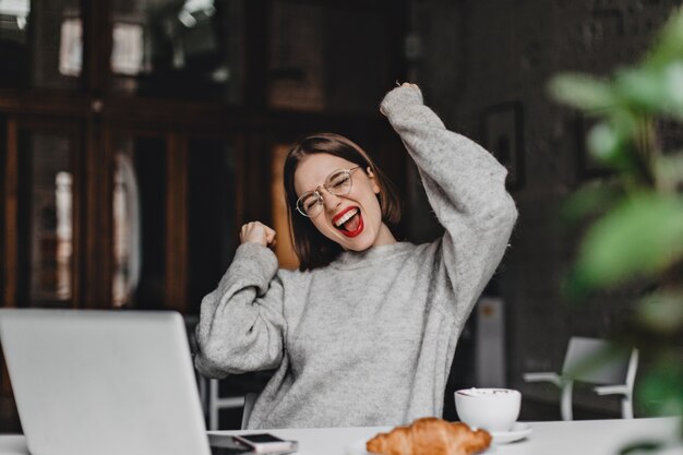 Une dame aux cheveux courts dans des verres avec du rouge à lèvres rouge exulte émotionnellement alors qu'elle est assise au lieu de travail avec un ordinateur portable et un croissant.