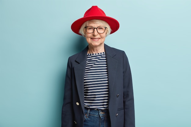 Dame aux cheveux gris à la mode avec le visage ridé, porte un chapeau élégant rouge, une veste et un jean, a un sourire agréable