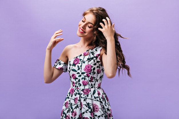 Dame aux cheveux longs en belle robe pose sur fond violet. Femme joyeuse bouclée dans des vêtements floraux mignon souriant sur fond isolé.