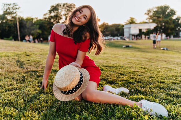 Dame aux cheveux noirs positive assise sur l'herbe et tenant un chapeau d'été. Élégante fille blanche avec plaisancier se détendre dans le parc.