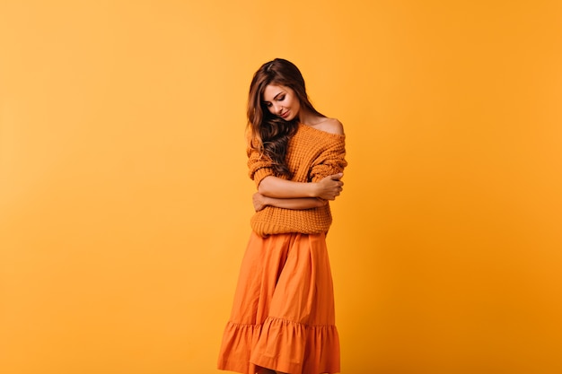 Dame aux cheveux noirs romantique en pull confortable posant sur jaune. Modèle féminin de bonne humeur en jupe orange regardant vers le bas.