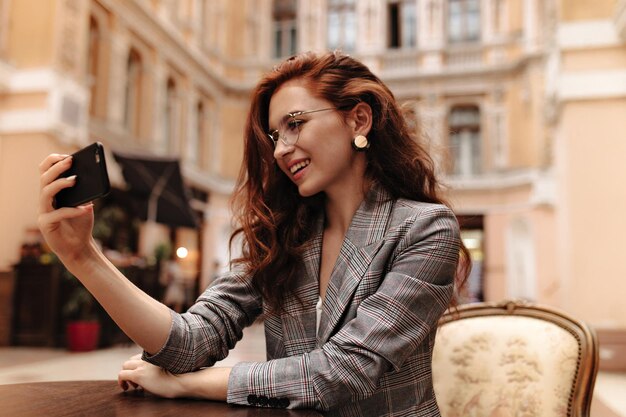 Une dame aux cheveux rouges en veste grise prend un selfie dans un café de rue