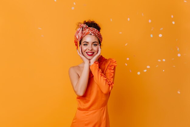 Dame aux yeux bleus en robe de soie orange et foulard touche doucement son visage et sourit sur fond orange avec des confettis