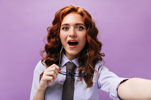 Une dame aux yeux verts tient des lunettes et prend un selfie sur un mur violet