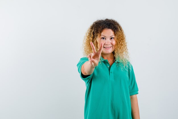 Dame blonde aux cheveux bouclés en T-shirt vert montrant le signe V et l'air joyeux, vue de face.