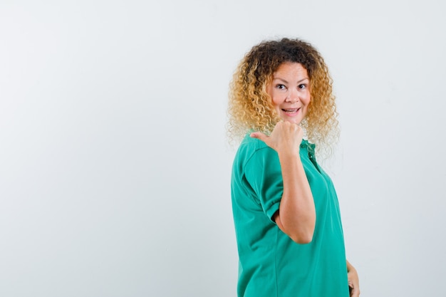 Dame Blonde Aux Cheveux Bouclés En T-shirt Vert Pointant De Côté Avec Le Pouce Et L'air Joyeux, Vue De Face.