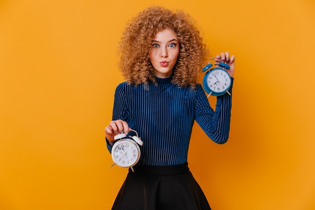 Photo gratuite dame blonde bouclée aux yeux bleus en pull rayé et jupe siffle et pose avec réveil sur l'espace jaune.