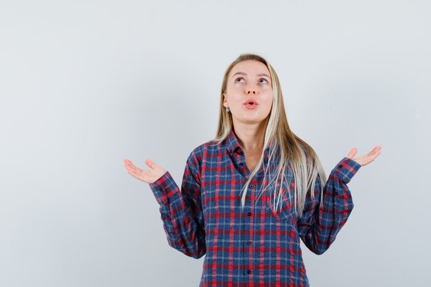 Dame blonde en chemise décontractée, levant les mains, regardant et regardant étonné, vue de face.