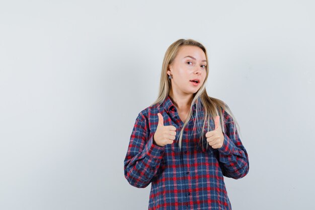 Dame blonde en chemise décontractée montrant les pouces vers le haut et l'air confiant, vue de face.