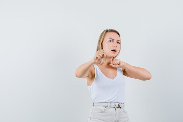 Dame blonde debout dans la lutte pose en maillot, pantalon et à la méchante, vue de face.