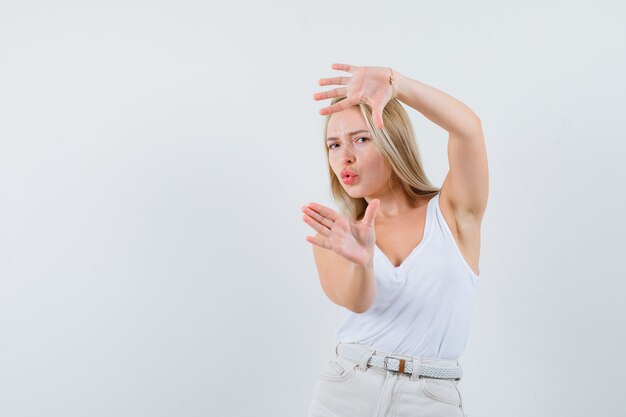 Dame blonde en maillot, pantalon faisant le geste du cadre et à la vue de face, confiant.