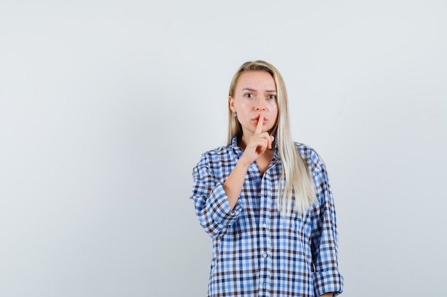 Dame blonde montrant le geste de silence en chemise à carreaux et à la troublé