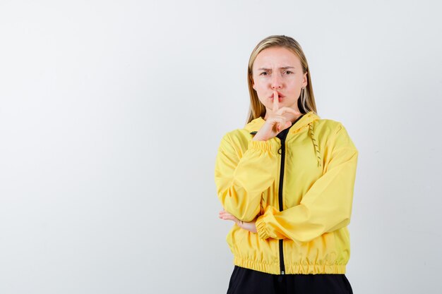 Dame blonde montrant le geste de silence en survêtement et à la recherche prudente. vue de face.