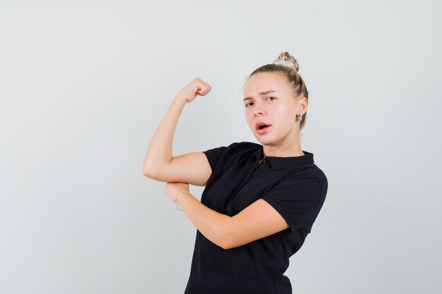 Dame blonde montrant les muscles du bras en t-shirt noir et l'air confiant, vue de face.