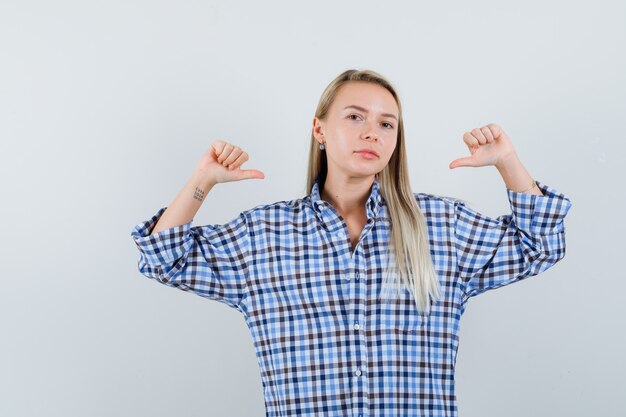 Dame blonde pointant sur elle-même avec les pouces en chemise à carreaux et à la confiance