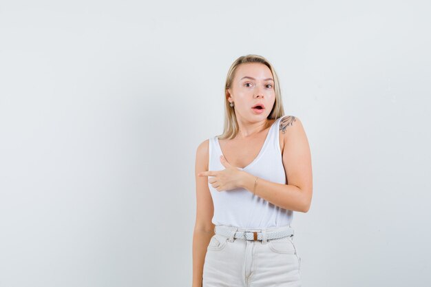 Dame blonde pointant vers le côté gauche en maillot, pantalon et à la perplexité, vue de face.