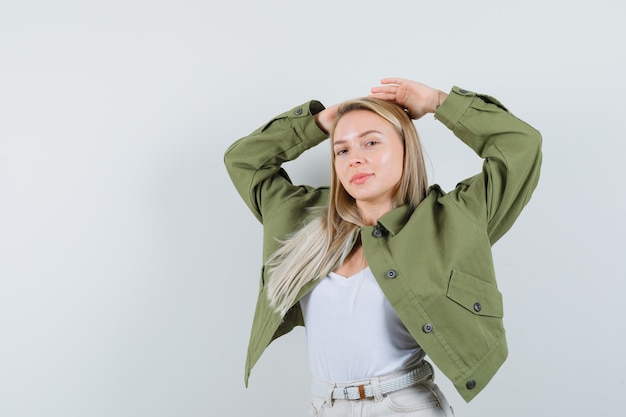 Dame blonde posant avec les mains derrière la tête en veste, pantalon et à la recherche attrayante, vue de face.