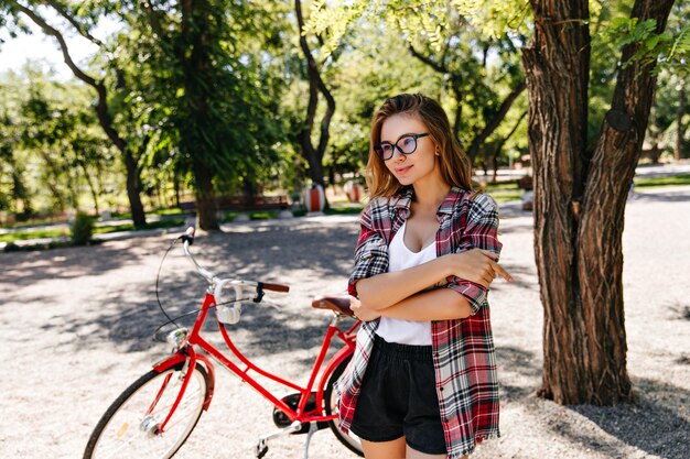 Dame blonde raffinée à lunettes posant après une balade à vélo. Portrait en plein air de fille debonair avec vélo rouge.