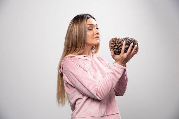 Une dame blonde en sweat-shirt rose fait un visage positif et heureux en tenant des cônes de chêne.