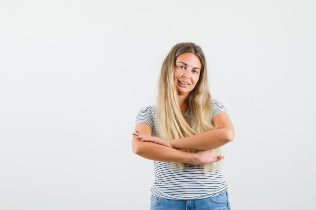 Dame blonde en t-shirt croisant les bras tout en souriant et à la coy, vue de face.