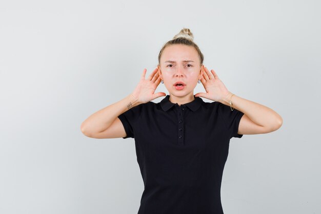 Dame blonde tenant les mains derrière les oreilles en t-shirt noir, vue de face.