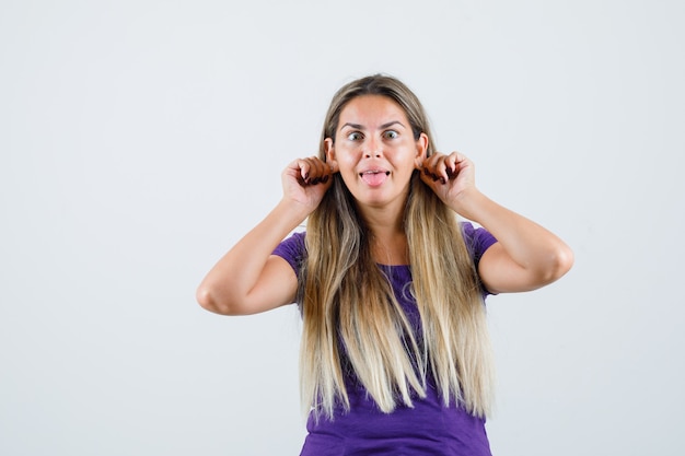 Dame blonde tirant vers le bas ses lobes d'oreille en t-shirt violet et à la drôle, vue de face.