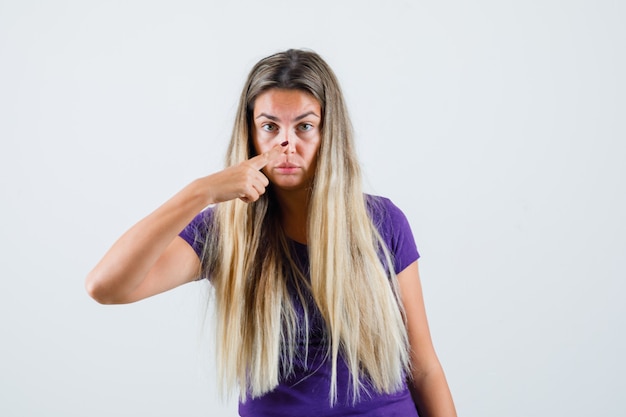 Dame blonde touchant le nez avec le doigt en t-shirt violet, vue de face.