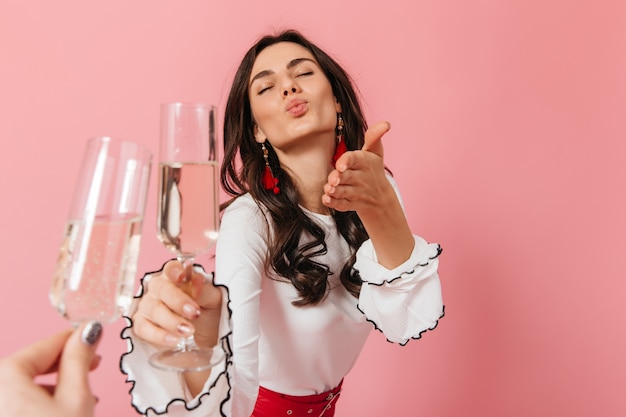 Dame avec des boucles d'oreilles rouges et un chemisier blanc envoie un baiser d'air et des verres de vin mousseux sur fond isolé.