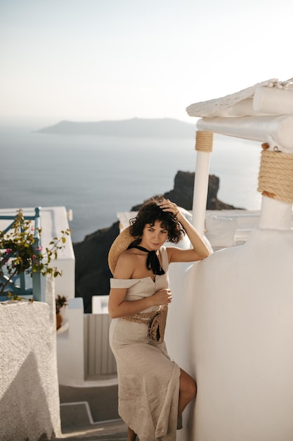 Dame brune en robe midi beige touche les cheveux Curly charmante femme en tenue élégante avec sac de paille pose dans la ville grecque avec vue sur la mer