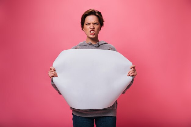 Dame en colère se penche sur la caméra et tient une feuille de papier avec place pour le texte. Femme émotionnelle en pantalon en denim à mal sur isolé