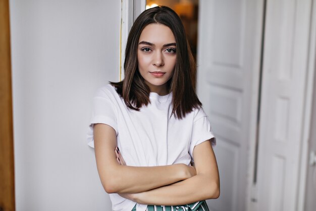 Dame confiante avec une coiffure droite debout dans son appartement, les bras croisés. Plan intérieur d'une femme européenne sérieuse porte un t-shirt blanc.