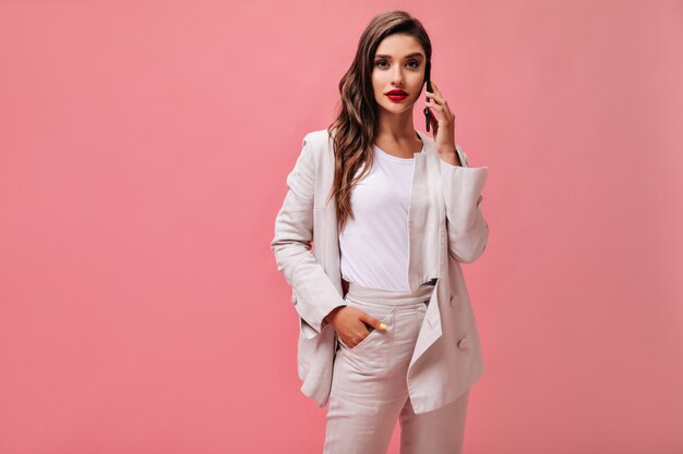 Dame en costume blanc se penche sur la caméra et parle au téléphone. Fille moderne en T-shirt léger et veste crème pose sur fond isolé.