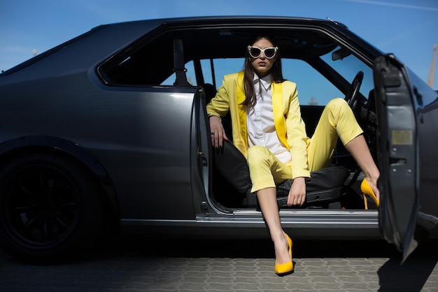 Photo gratuite dame élégante assise dans la voiture avec porte ouverte. fashion girl au volant d'une voiture en costume. fille chic dans une voiture de sport.