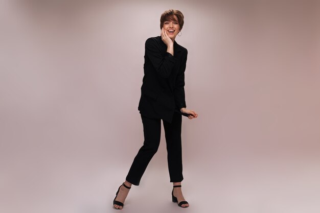 Dame élégante en costume riant sur fond isolé. Portrait de femme vêtue d'une veste noire et d'un pantalon posant et souriant sur fond blanc