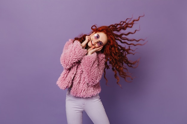 Dame élégante en manteau en peau de mouton, pantalon skinny souriant et jouant les cheveux sur l'espace lilas.