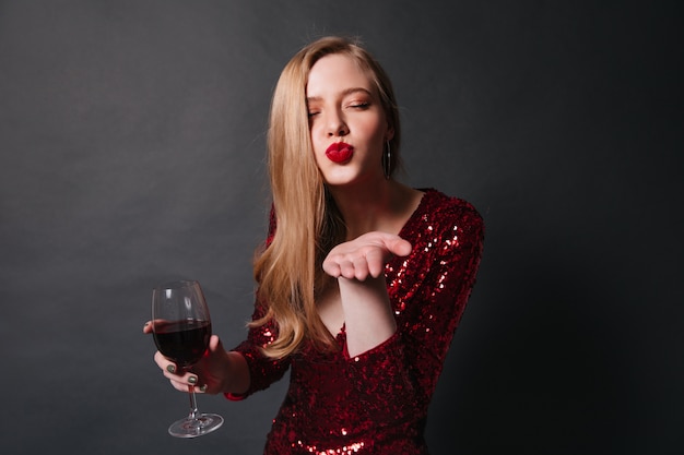 Dame élégante avec verre à vin envoyant un baiser d'air. Photo de Studio d'une fille blonde en robe rouge, boire du vin à la fête.