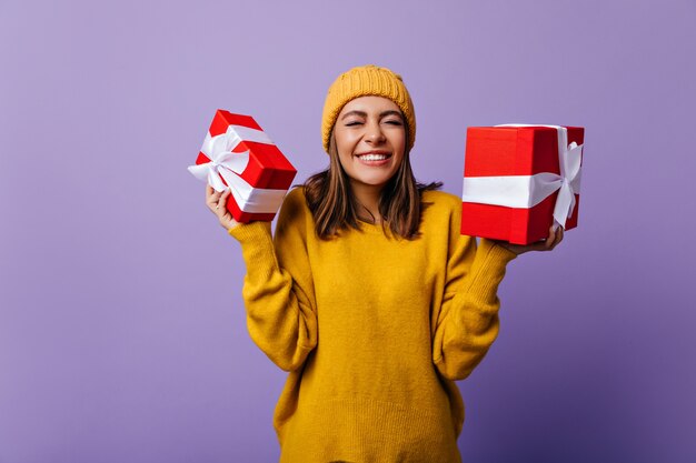 Dame enthousiaste en bonnet tricoté posant avec des cadeaux de Noël. Joyeuse fille d'anniversaire bénéficiant de cadeaux.