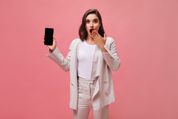 Dame étonnée en costume montre le téléphone sur fond rose. Une brune surprise en tenue beige élégante tient le smartphone et pose pour la caméra.