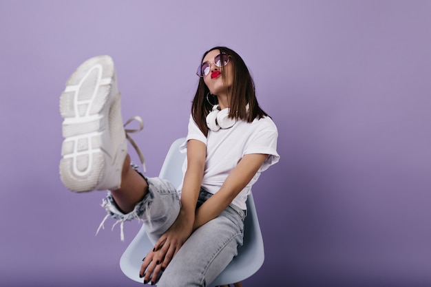 Photo gratuite dame européenne confiante en baskets blanches assis sur une chaise et faisant des grimaces. portrait de fille merveilleuse posant.