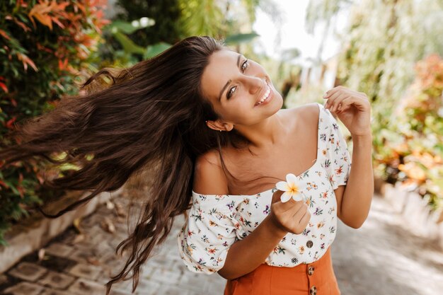 Dame avec une fleur blanche dans sa main joue avec les cheveux Femme en tenue d'été lumineuse avec sourire posant sur fond de plantes