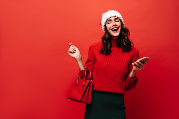 Photo gratuite dame gaie en pull et jupe verte riant sur fond rouge fille aux cheveux ondulés en casquette blanche avec sac à main tenant le téléphone sur fond isolé