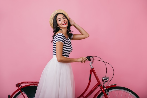 Photo gratuite dame heureuse bronzée au chapeau s'amusant à vélo. portrait intérieur de fille de bonne humeur en jupe luxuriante debout.