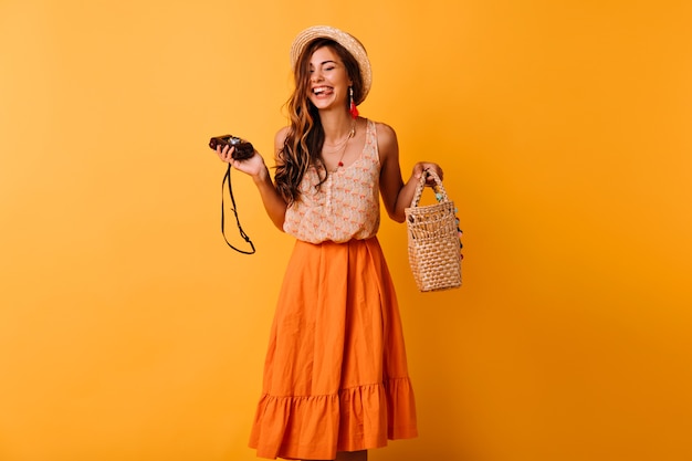 Dame heureuse dans des vêtements d'été à la mode posant avec caméra sur jaune. Positive belle fille au chapeau refroidissant en studio.